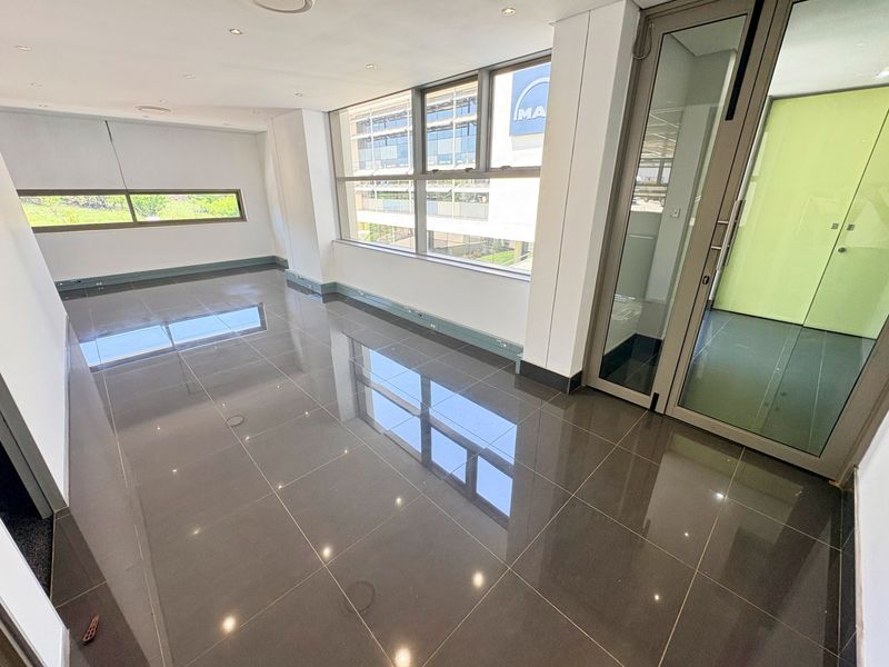 A bright, modern, and empty office space featuring polished tiled floors, large windows providing natural light, and a sleek glass partition door.