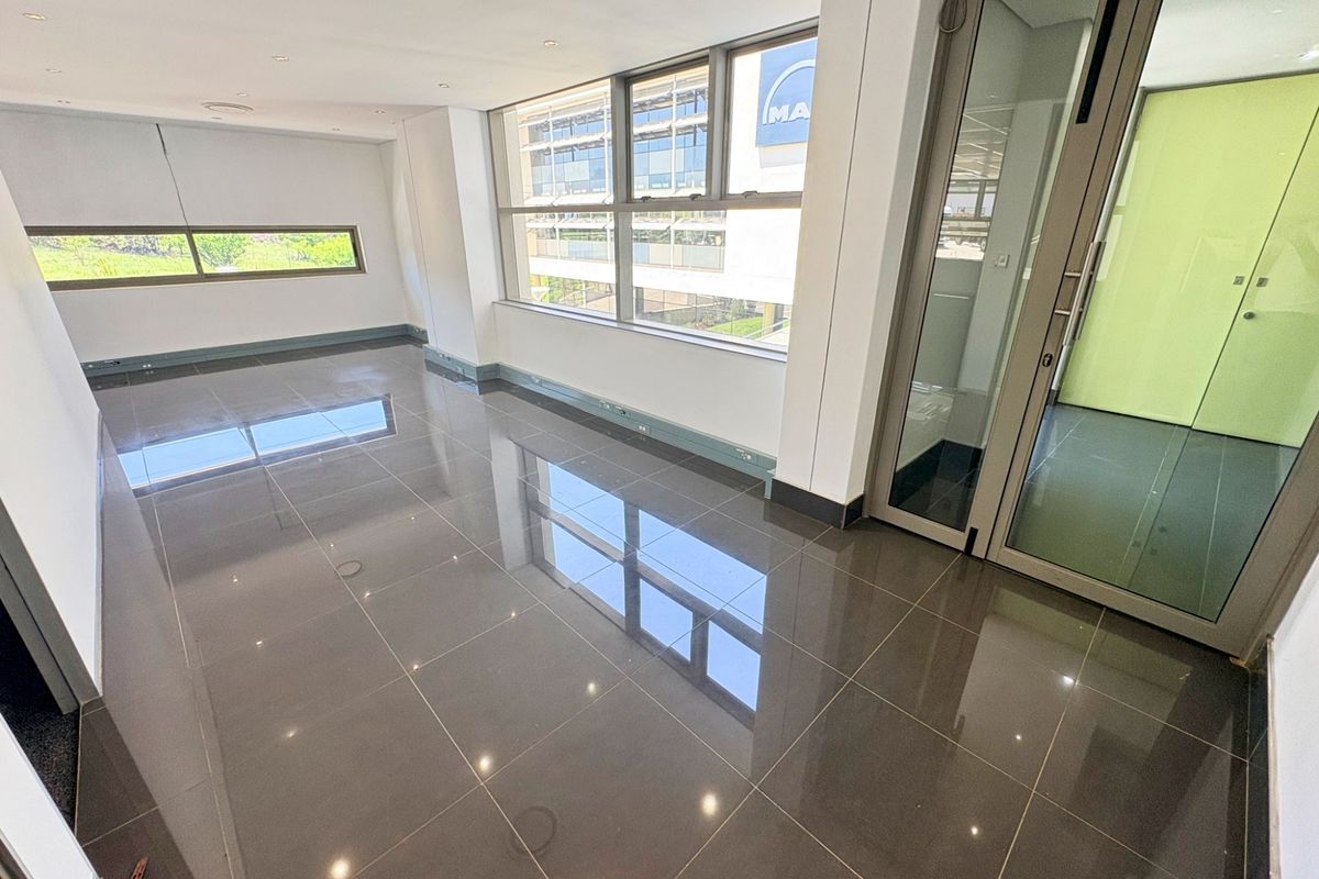 A bright, modern, and empty office space featuring polished tiled floors, large windows providing natural light, and a sleek glass partition door.