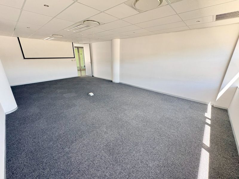 A spacious, empty office area featuring a suspended ceiling, recessed lighting, carpeted flooring, and a wall-mounted projector screen.