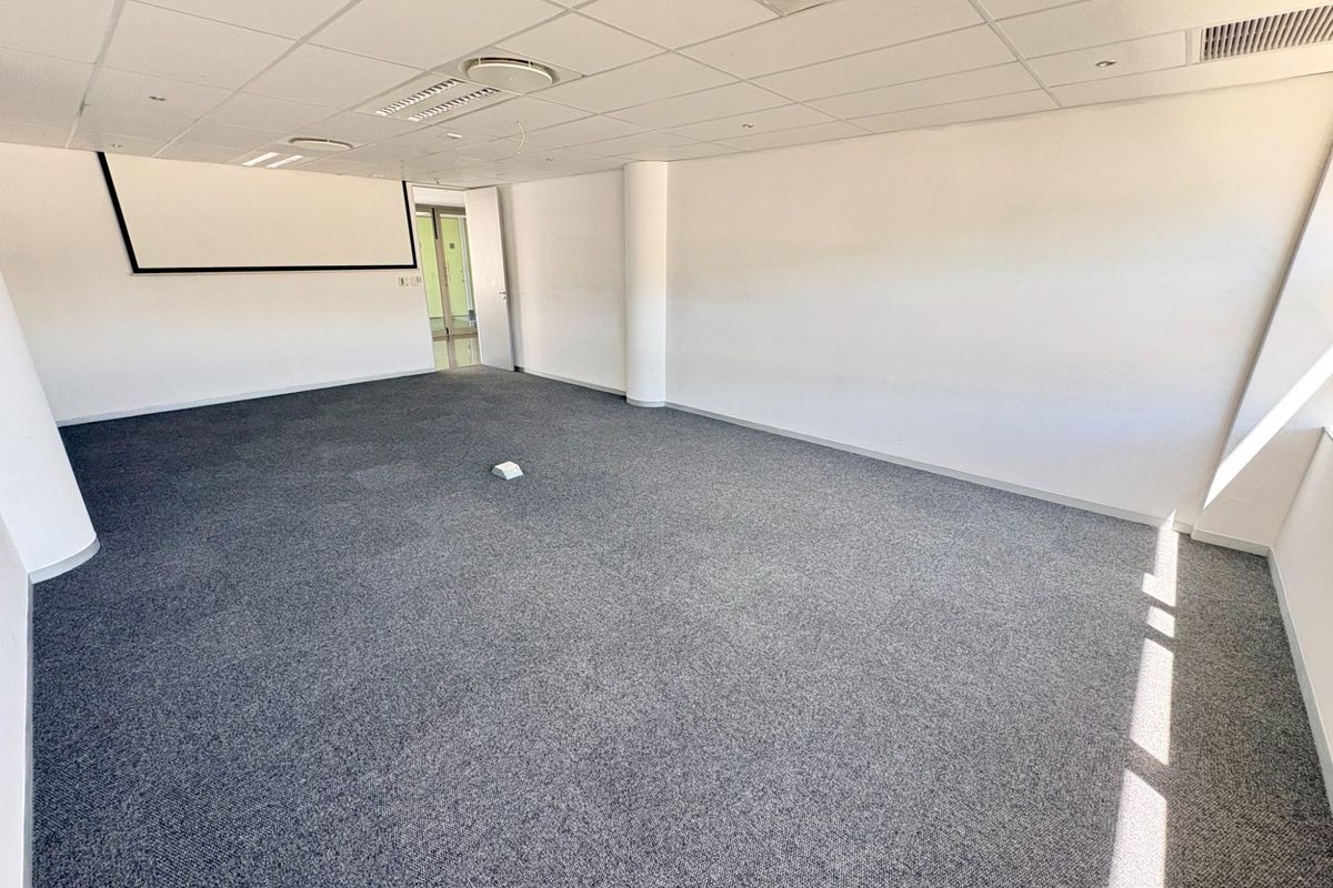 A spacious, empty office area featuring a suspended ceiling, recessed lighting, carpeted flooring, and a wall-mounted projector screen.