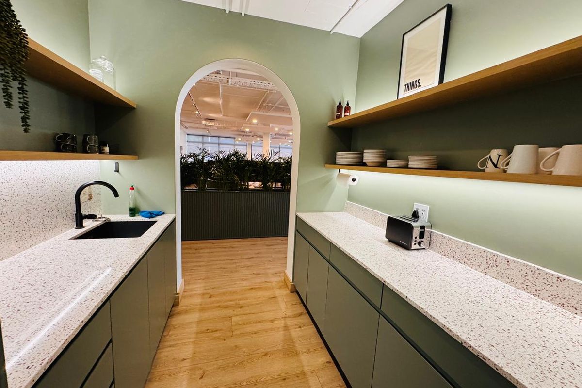 A modern office kitchenette featuring sage green cabinetry, terrazzo countertops, floating shelves with integrated lighting, and a stylish arched entryway.