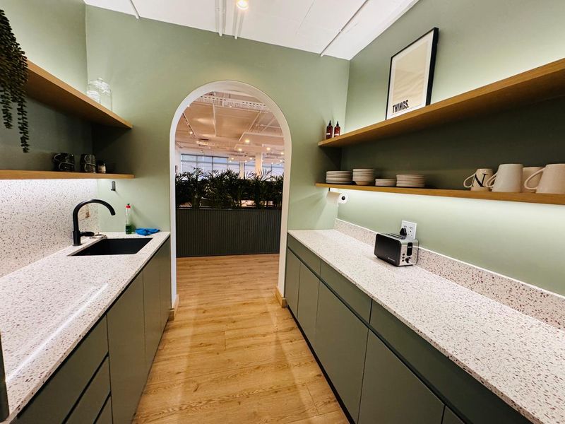 A modern office kitchenette featuring sage green cabinetry, terrazzo countertops, floating shelves with integrated lighting, and a stylish arched entryway.