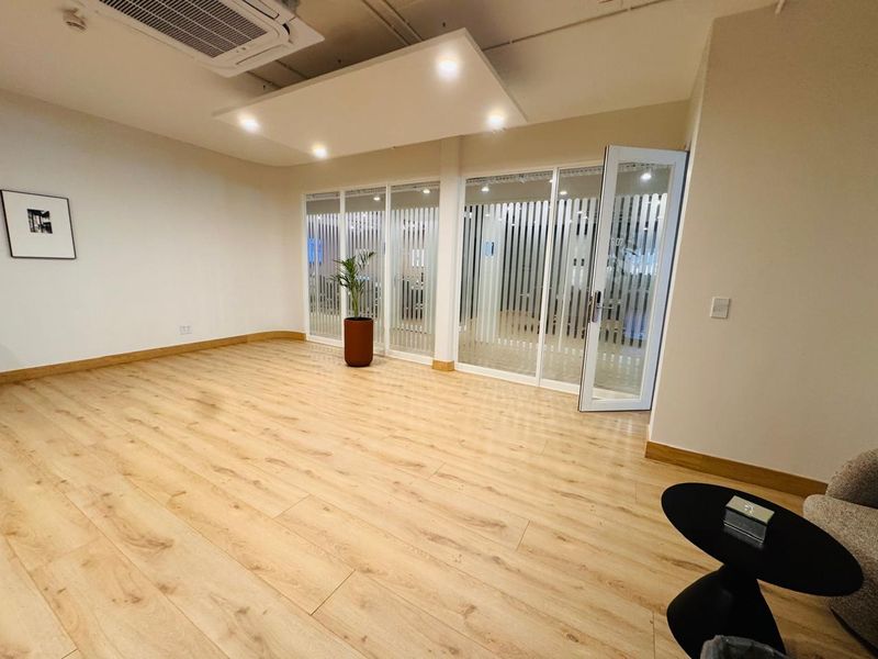 A modern, bright office space featuring light wood-look laminate flooring, glass partition walls, and integrated ceiling lighting with ducted air conditioning.