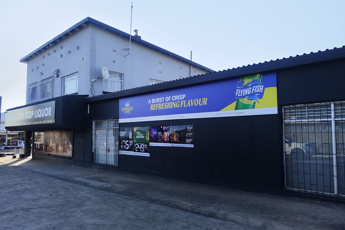 Front exterior view of a commercial liquor store building featuring prominent signage, security bars on windows, and a corrugated metal roof.