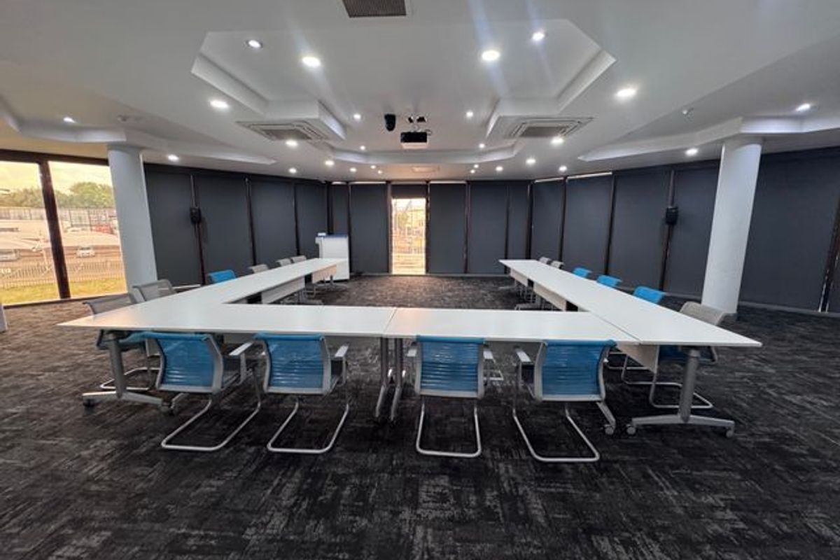 A professional boardroom or meeting space featuring recessed ceiling lighting, ducted air conditioning, and large windows with dark roller blinds.