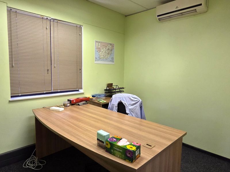 A functional office space featuring light green walls, carpeted flooring, a window with venetian blinds, and a wall-mounted air conditioning unit.