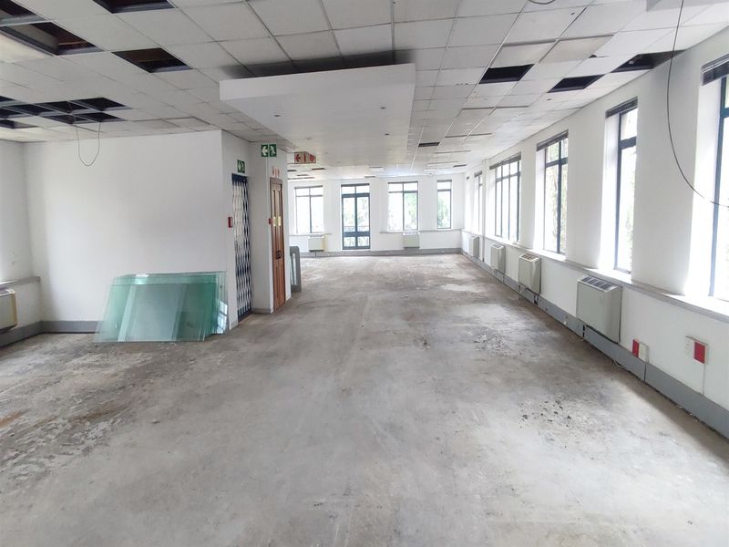 A large, vacant open-plan office space featuring a suspended ceiling with missing tiles, large windows, and HVAC units, requiring significant refurbishment.
