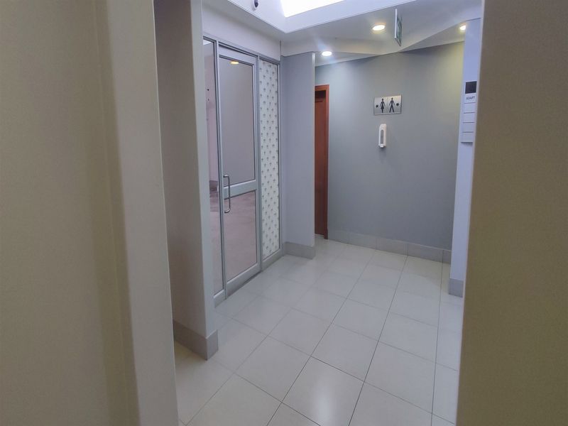 A clean, well-lit office hallway featuring tiled flooring, a skylight, and access to restroom facilities.