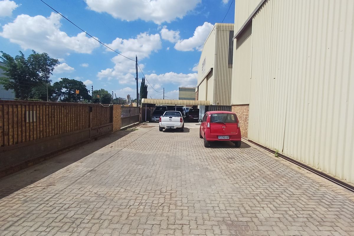 Exterior view of an industrial property featuring a paved driveway, perimeter brick wall, and metal-clad warehouse structure with covered parking area.
