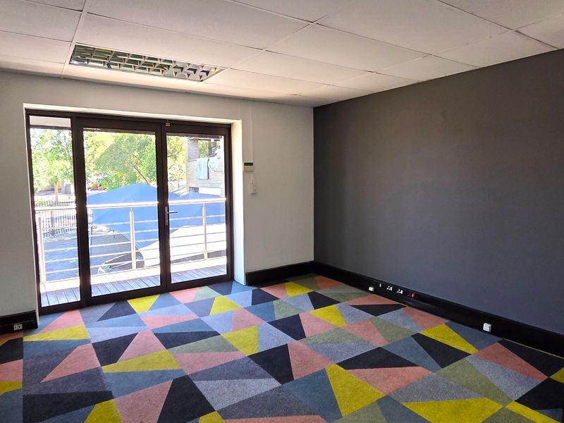 A bright, empty office space featuring patterned carpet tiles, a drop ceiling with recessed lighting, and direct access to a private balcony.