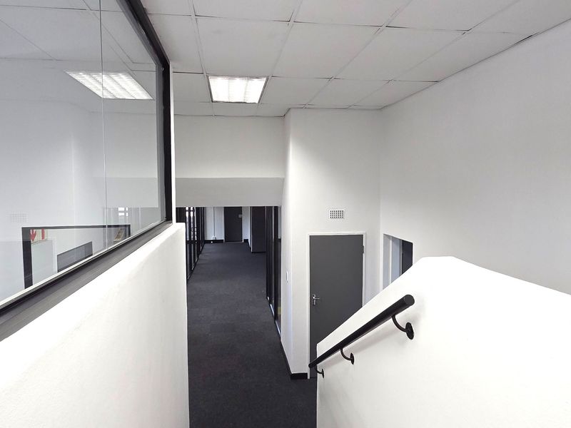 A clean, modern office hallway featuring a staircase with a black handrail, glass partitioning, and neutral carpet flooring.