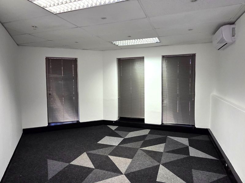 A clean, empty office space featuring modern geometric carpet tiles, suspended ceiling with recessed lighting, and a wall-mounted air conditioning unit.