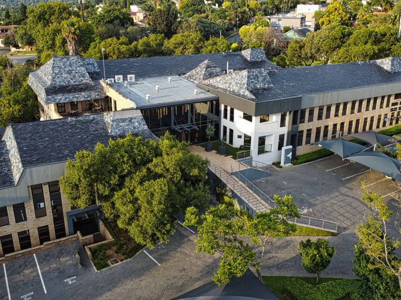 Aerial view of a modern office complex featuring a slate roof, glass facade, brick exterior, and extensive paved parking with shade cloth carports.