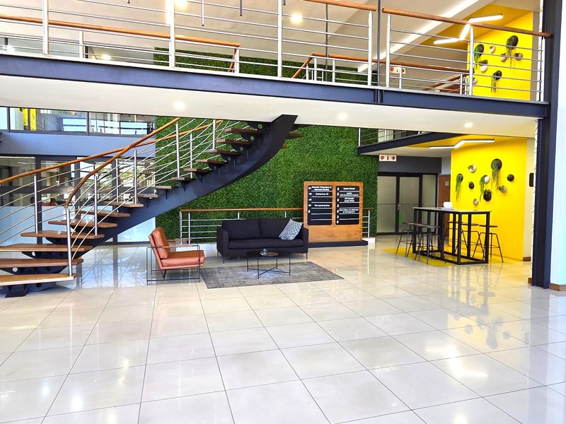 Modern open-plan office lobby featuring a striking floating staircase, mezzanine level, polished tile flooring, and a vibrant green feature wall.