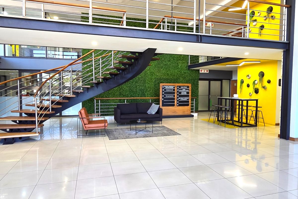 Modern open-plan office lobby featuring a striking floating staircase, mezzanine level, polished tile flooring, and a vibrant green feature wall.
