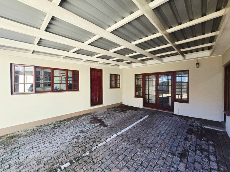 A covered outdoor carport area featuring paved flooring and timber-framed glass doors providing access to the office building.