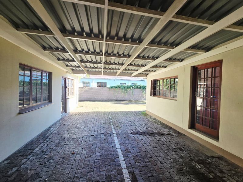 A covered outdoor parking or utility area featuring a corrugated metal roof, timber-framed windows and doors, and a brick-paved surface.