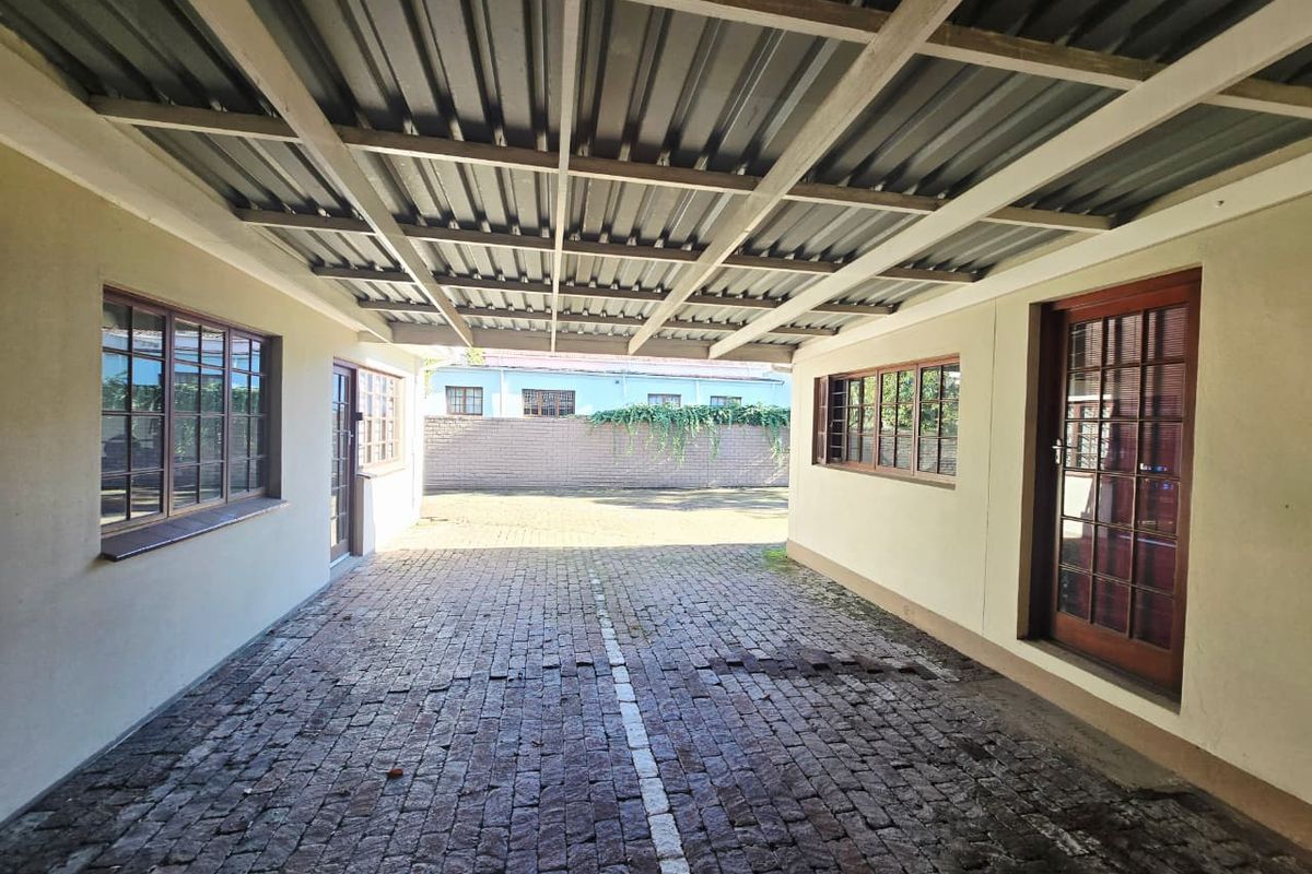 A covered outdoor parking or utility area featuring a corrugated metal roof, timber-framed windows and doors, and a brick-paved surface.