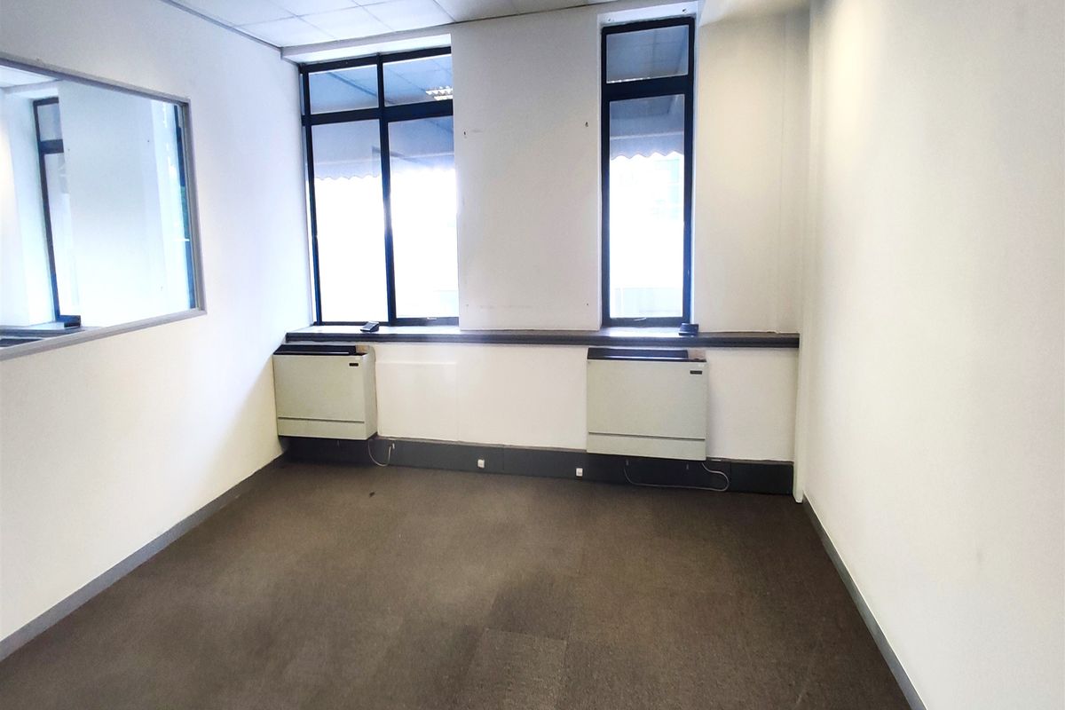 An empty office space featuring carpet flooring, suspended ceilings with recessed lighting, and two HVAC units positioned beneath large windows.