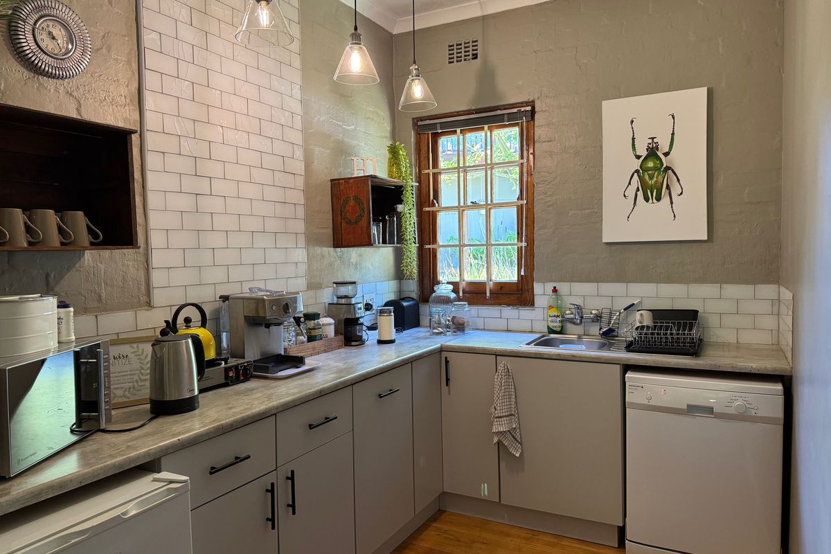 A functional office kitchenette featuring grey cabinetry, stone-look countertops, subway tile splashbacks, a stainless steel sink, and timber flooring.