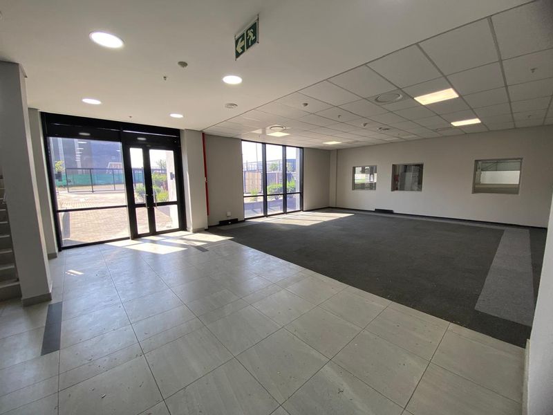 Spacious open-plan industrial office area featuring a mix of tiled and carpeted flooring, large windows for natural light, and a suspended ceiling.