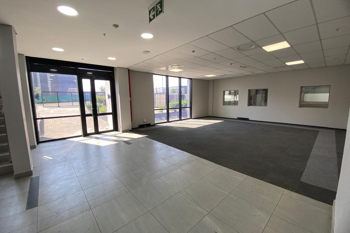 Spacious open-plan industrial office area featuring a mix of tiled and carpeted flooring, large windows for natural light, and a suspended ceiling.