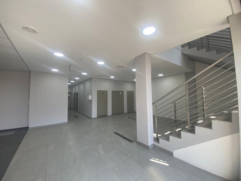 A spacious, modern industrial hallway featuring tiled flooring, recessed ceiling lighting, and a stainless steel staircase railing leading to the upper level.
