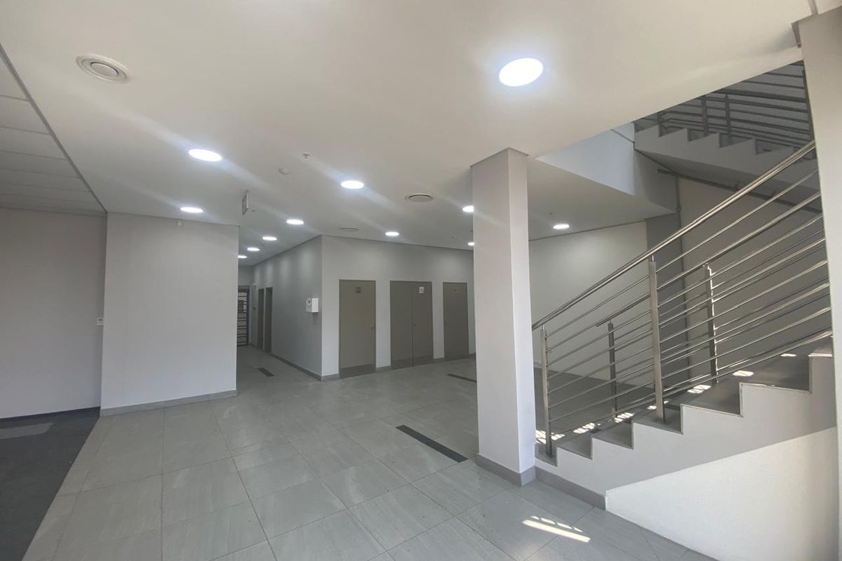 A spacious, modern industrial hallway featuring tiled flooring, recessed ceiling lighting, and a stainless steel staircase railing leading to the upper level.