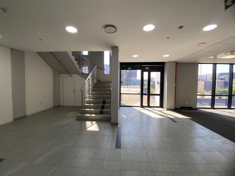 Spacious industrial entrance lobby featuring tiled flooring, recessed lighting, and a central staircase with metal railings, leading to a glass-paneled exterior entrance.