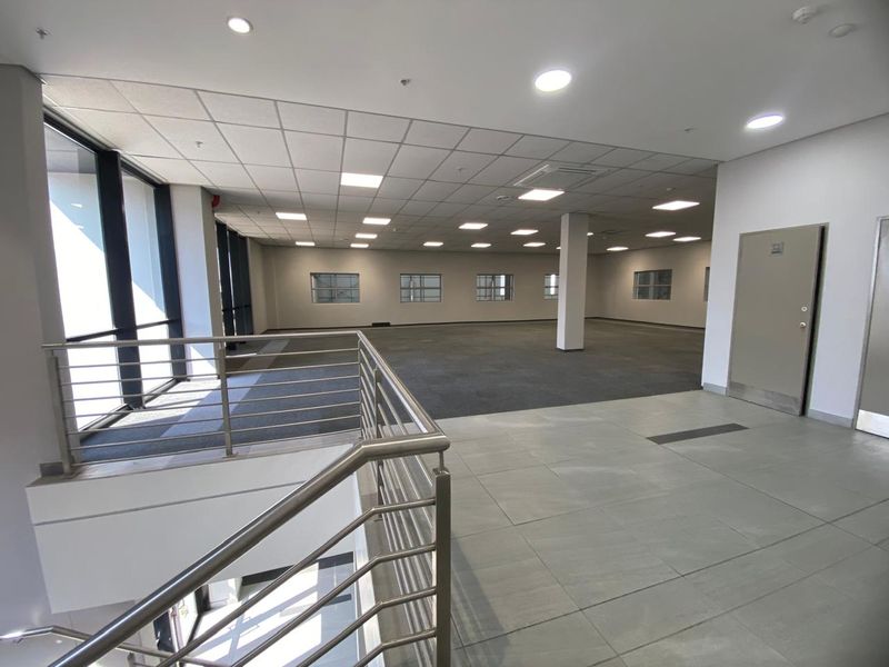 Spacious open-plan industrial office area featuring a mix of carpet and tile flooring, large windows for natural light, and a modern stainless steel balustrade.