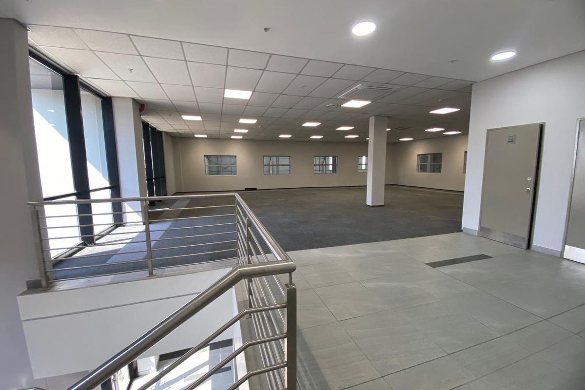 Spacious open-plan industrial office area featuring a mix of carpet and tile flooring, large windows for natural light, and a modern stainless steel balustrade.