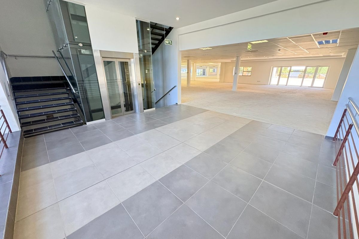 Spacious office entrance lobby featuring modern tiled flooring, a glass elevator, and an open-plan layout leading into a bright, vacant workspace.