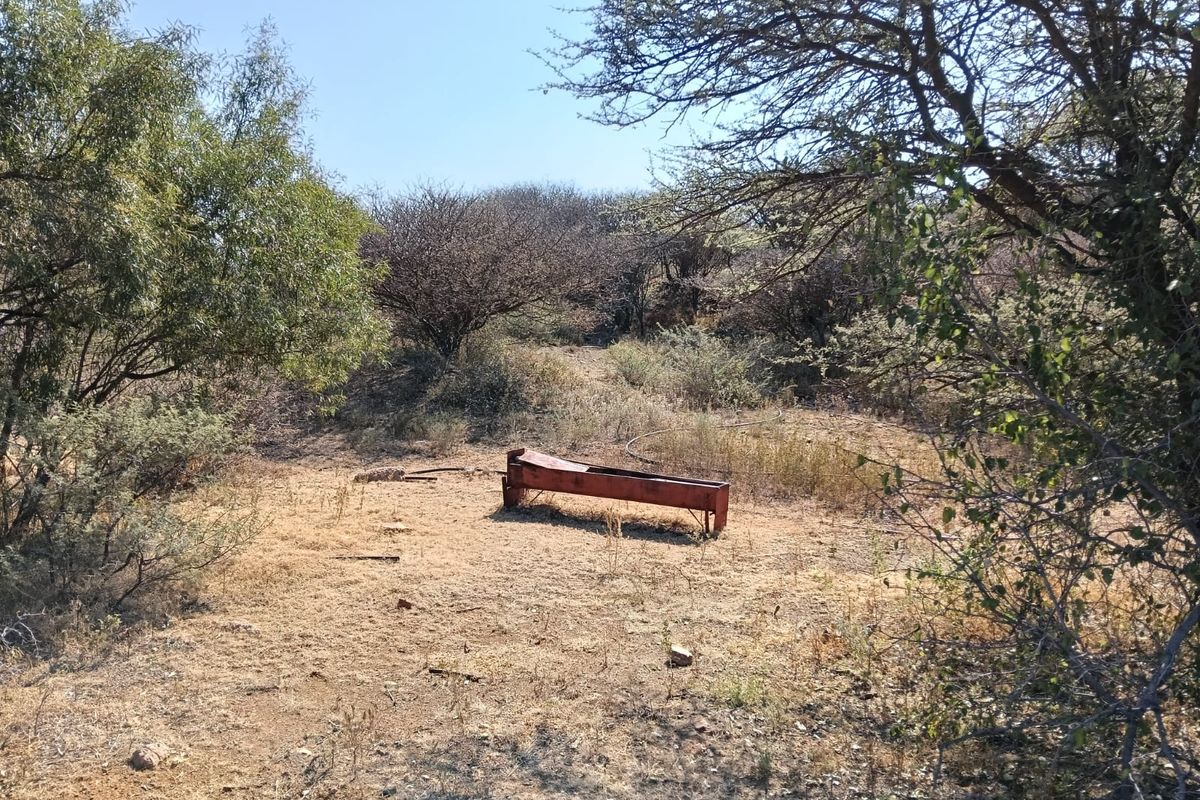 A rural farm landscape featuring a weathered metal livestock water trough situated in a dry, open field surrounded by native trees and shrubs.