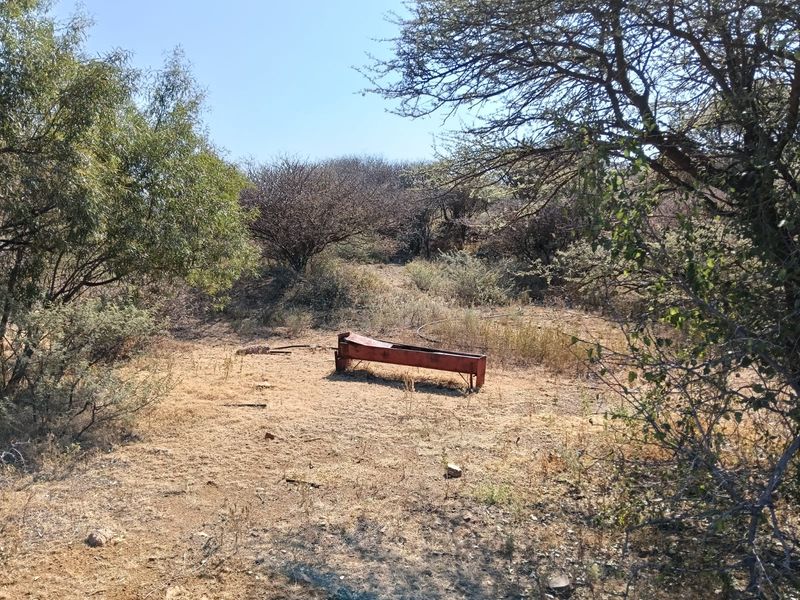 A rural farm landscape featuring a weathered metal livestock water trough situated in a dry, open field surrounded by native trees and shrubs.