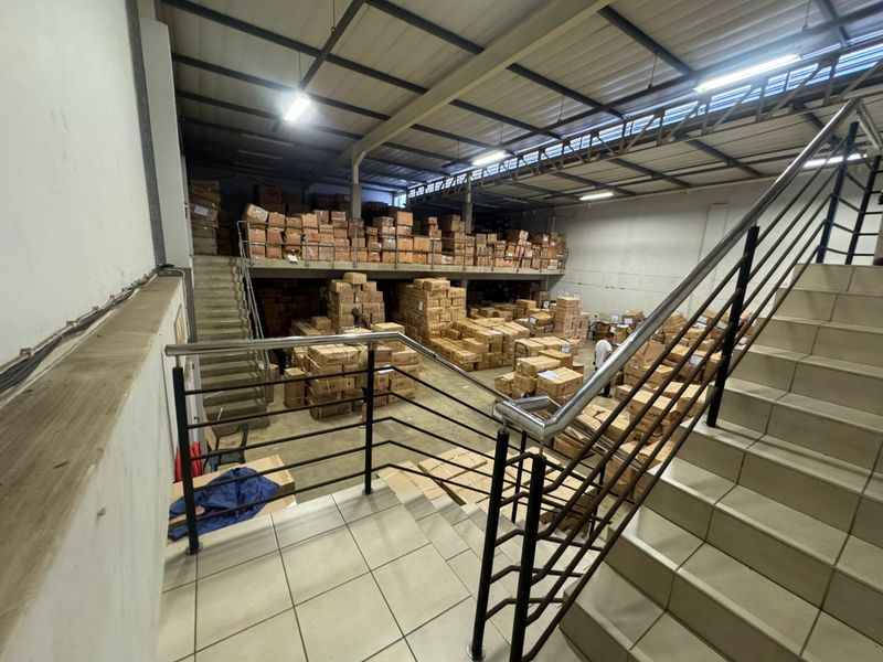 Industrial warehouse space featuring a mezzanine level, steel staircase, and high-clearance exposed roof trusses, suitable for storage or logistics operations.