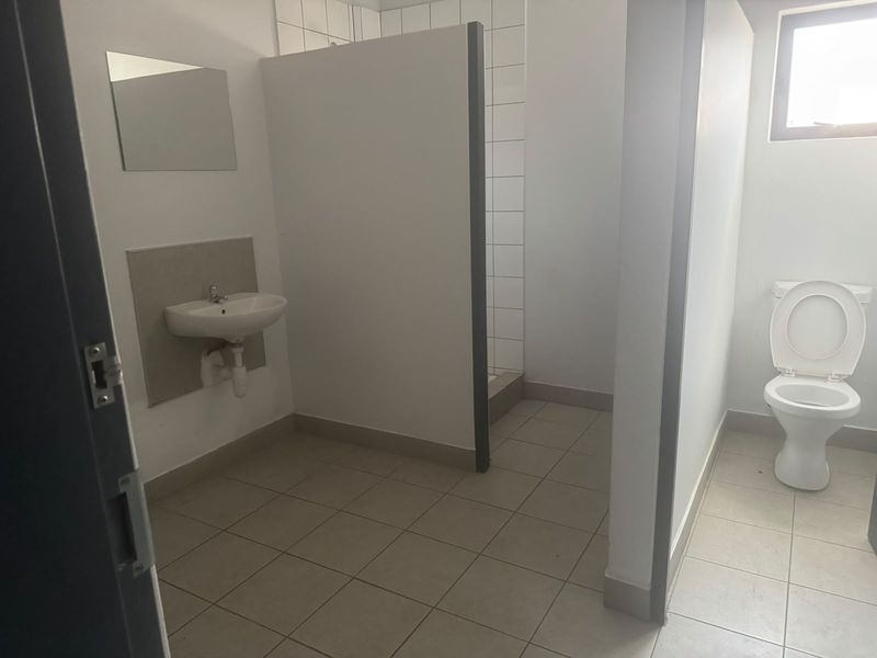 Functional industrial bathroom featuring a wall-mounted basin, a tiled shower cubicle, and a standard flush toilet with neutral tiled flooring.