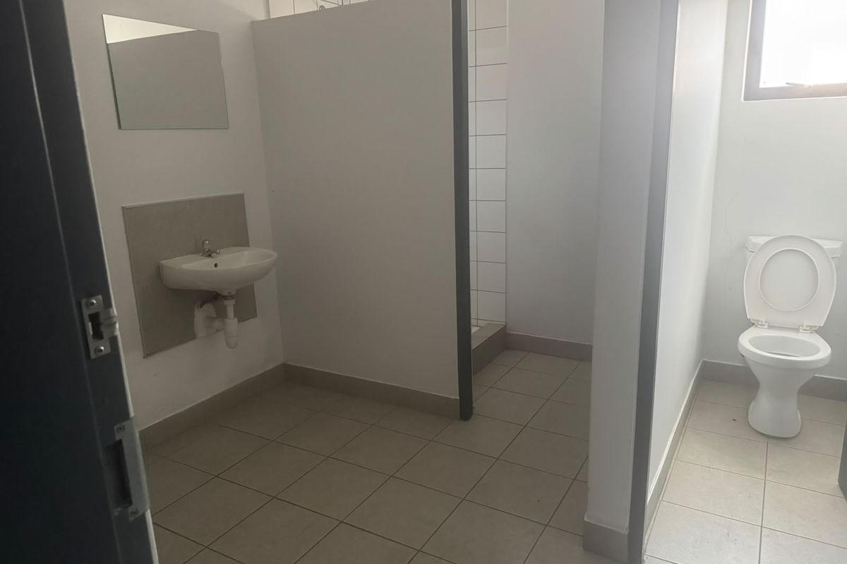 Functional industrial bathroom featuring a wall-mounted basin, a tiled shower cubicle, and a standard flush toilet with neutral tiled flooring.