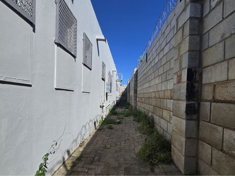 A narrow exterior side passage featuring a paved walkway, security-fortified boundary walls with razor wire, and metal security window grilles.