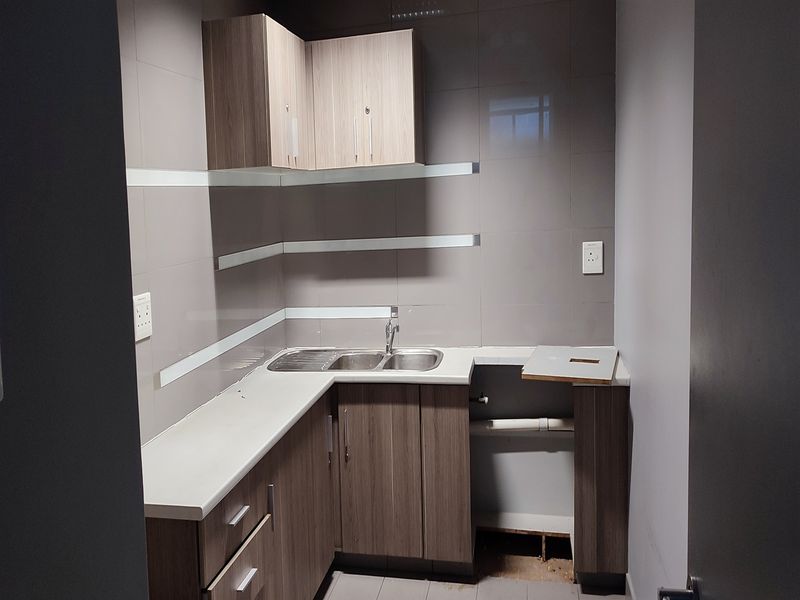 A small kitchenette area featuring wood-finish cabinetry, a stainless steel sink, and tiled walls, currently requiring minor repairs to the base cabinetry.