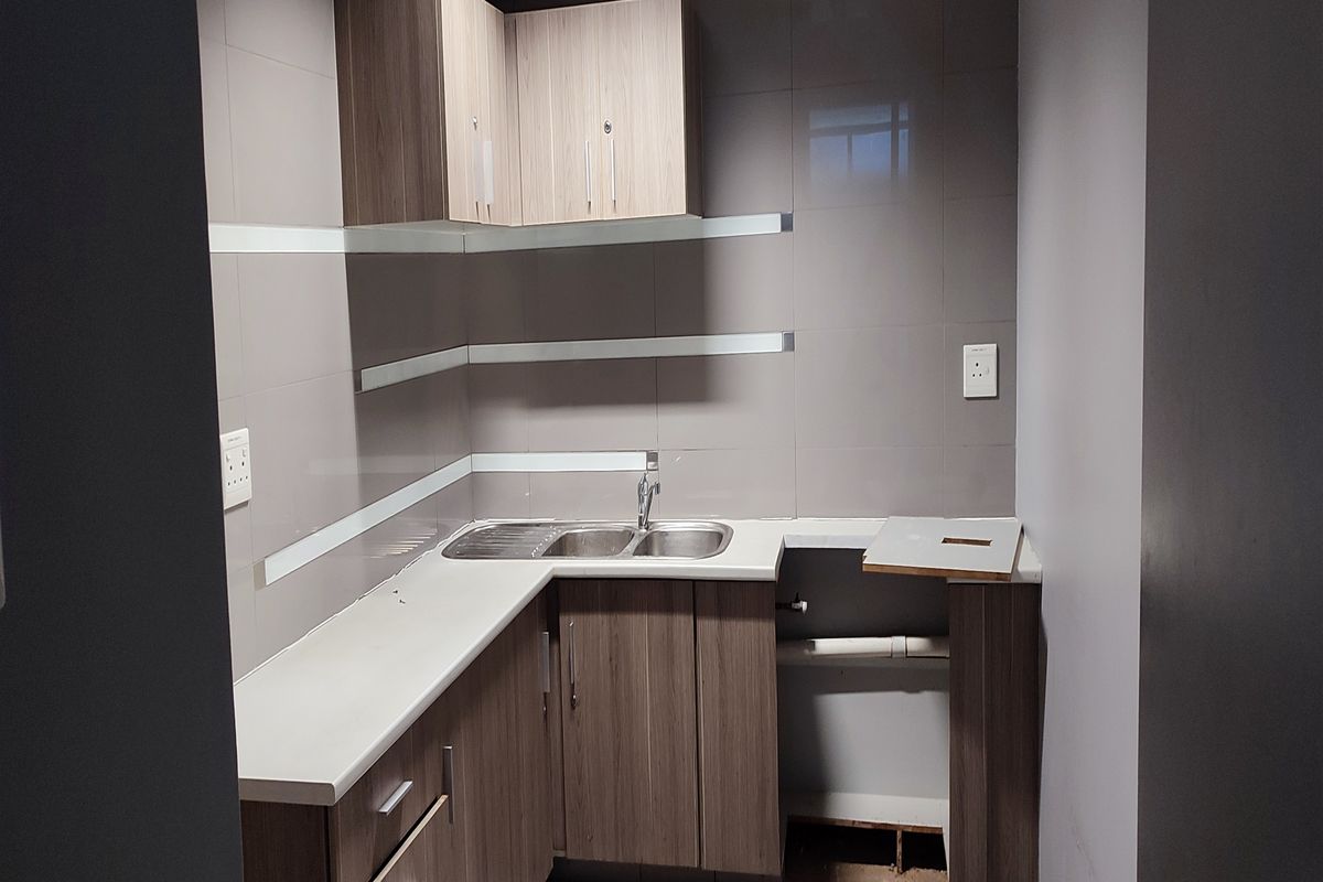 A small kitchenette area featuring wood-finish cabinetry, a stainless steel sink, and tiled walls, currently requiring minor repairs to the base cabinetry.