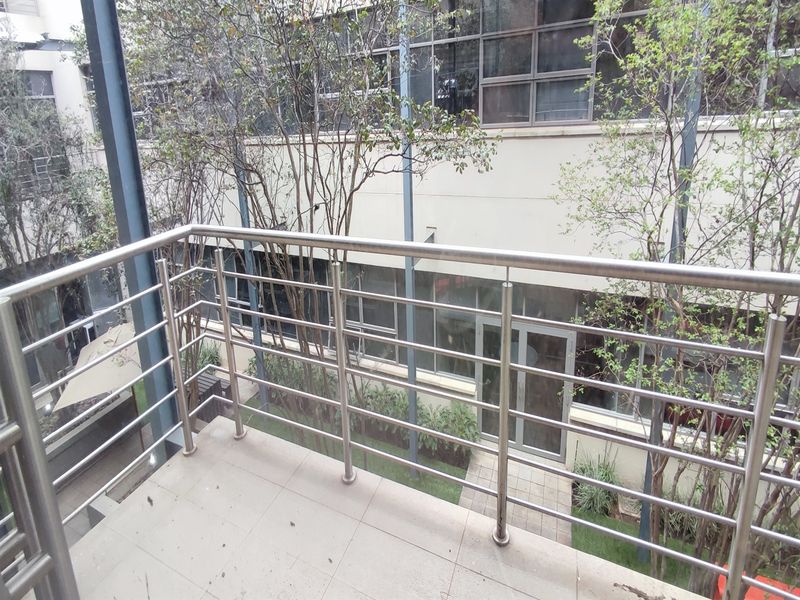 A clean, elevated balcony featuring a modern stainless steel balustrade and tiled flooring, overlooking a landscaped courtyard area with large office windows.