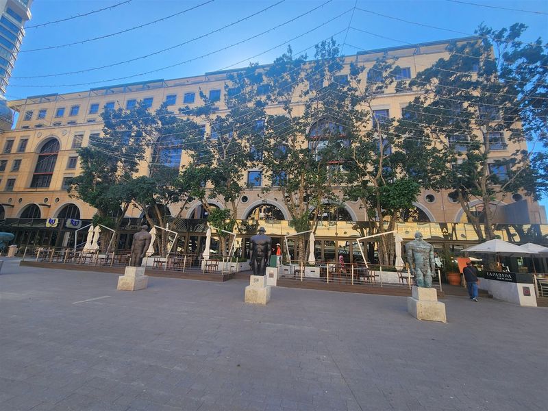 A wide-angle view of a multi-story office building facade featuring an expansive paved plaza, mature trees, and an integrated outdoor dining deck.