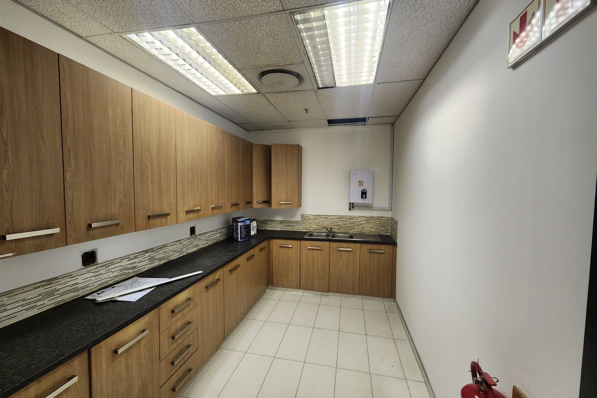A functional office kitchenette featuring wood-finish cabinetry, dark laminate countertops, a stainless steel sink, and tiled backsplash under a drop ceiling.