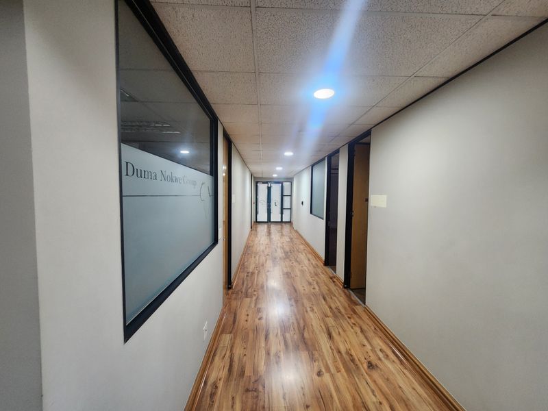 A clean, well-lit office hallway featuring wood-look laminate flooring, a suspended ceiling with recessed lighting, and glass-partitioned office entrances.