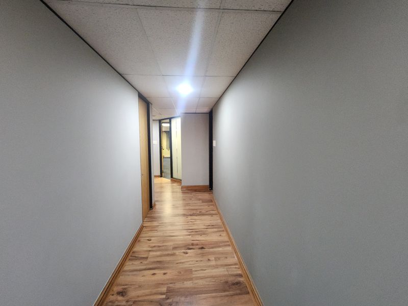 A clean, well-maintained office hallway featuring modern laminate flooring, neutral grey walls, and a suspended ceiling with recessed lighting.