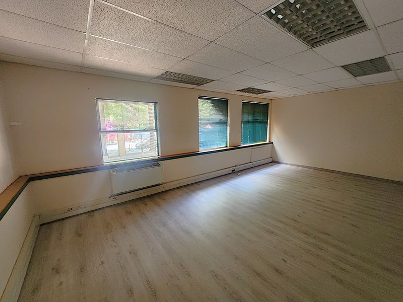 A vacant office space featuring laminate flooring, a suspended ceiling with recessed lighting, and wall-mounted HVAC units with large windows providing natural light.