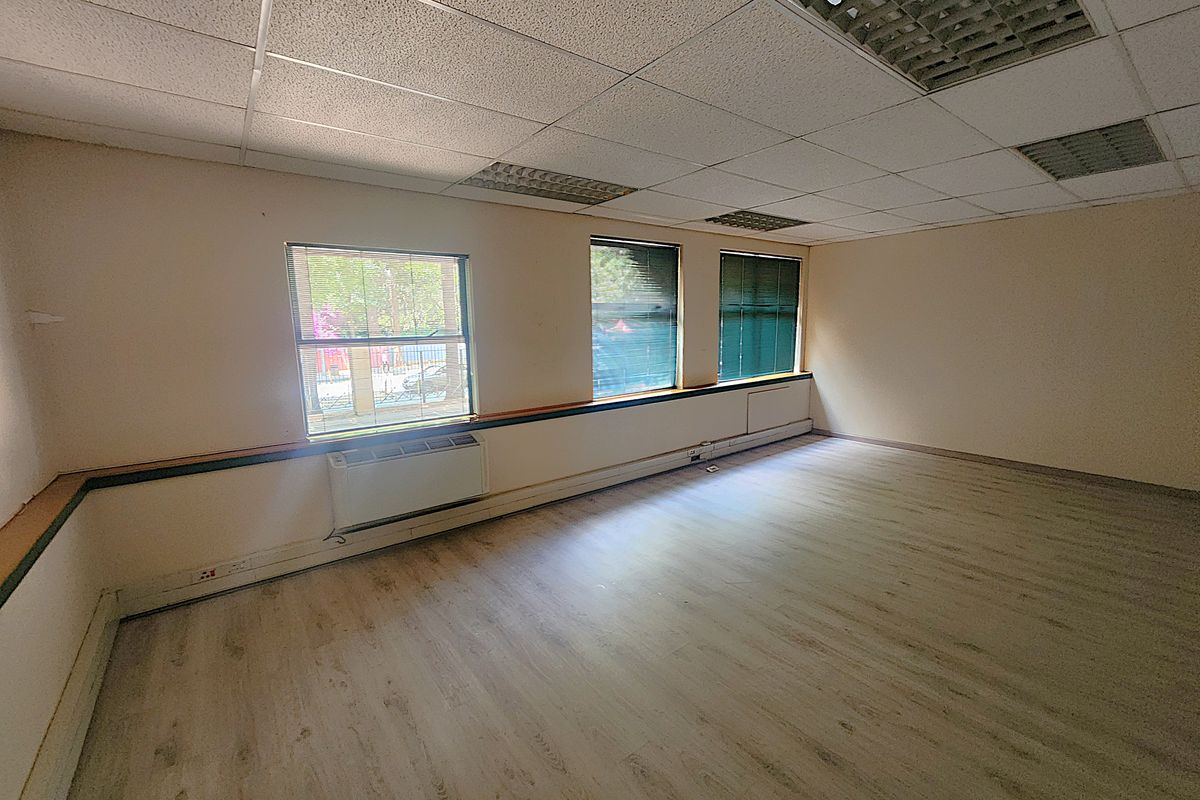 A vacant office space featuring laminate flooring, a suspended ceiling with recessed lighting, and wall-mounted HVAC units with large windows providing natural light.