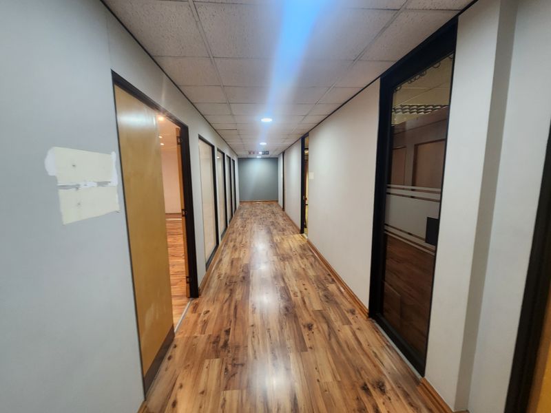 A long office hallway featuring wood-look laminate flooring, a suspended ceiling with recessed lighting, and multiple glass-paneled office doors.