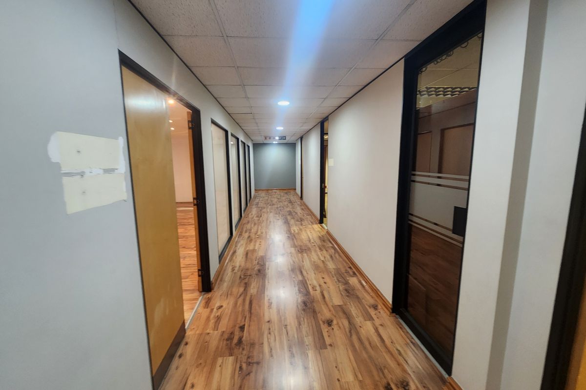 A long office hallway featuring wood-look laminate flooring, a suspended ceiling with recessed lighting, and multiple glass-paneled office doors.
