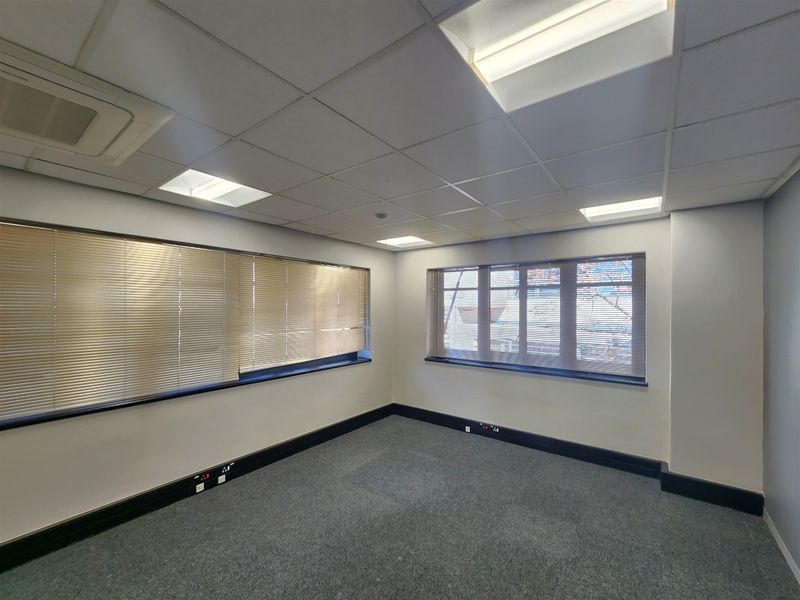 A clean, empty office space featuring a suspended ceiling with recessed lighting, air conditioning, carpeted flooring, and large windows with fitted venetian blinds.
